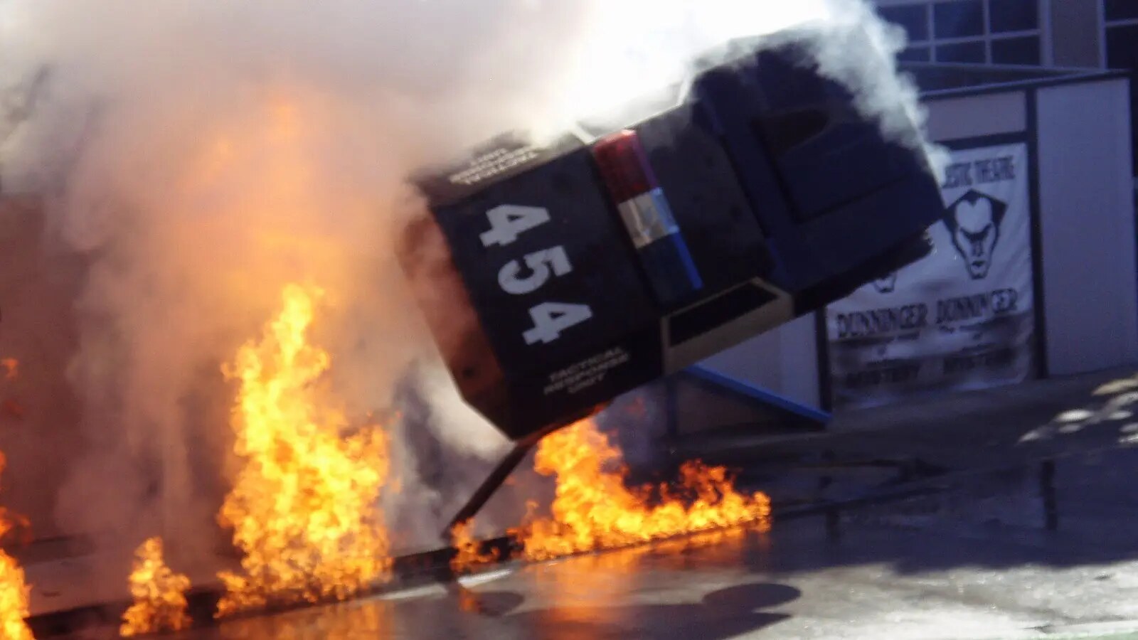 A full-size police-show vehicle flips sideways above a line of fire as smoke fills the stunt arena.