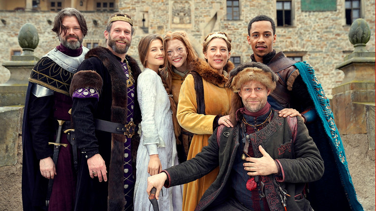 Seven cast members of The Witch Princess pose in costume in front of a castle courtyard in the official ZDF ensemble still.