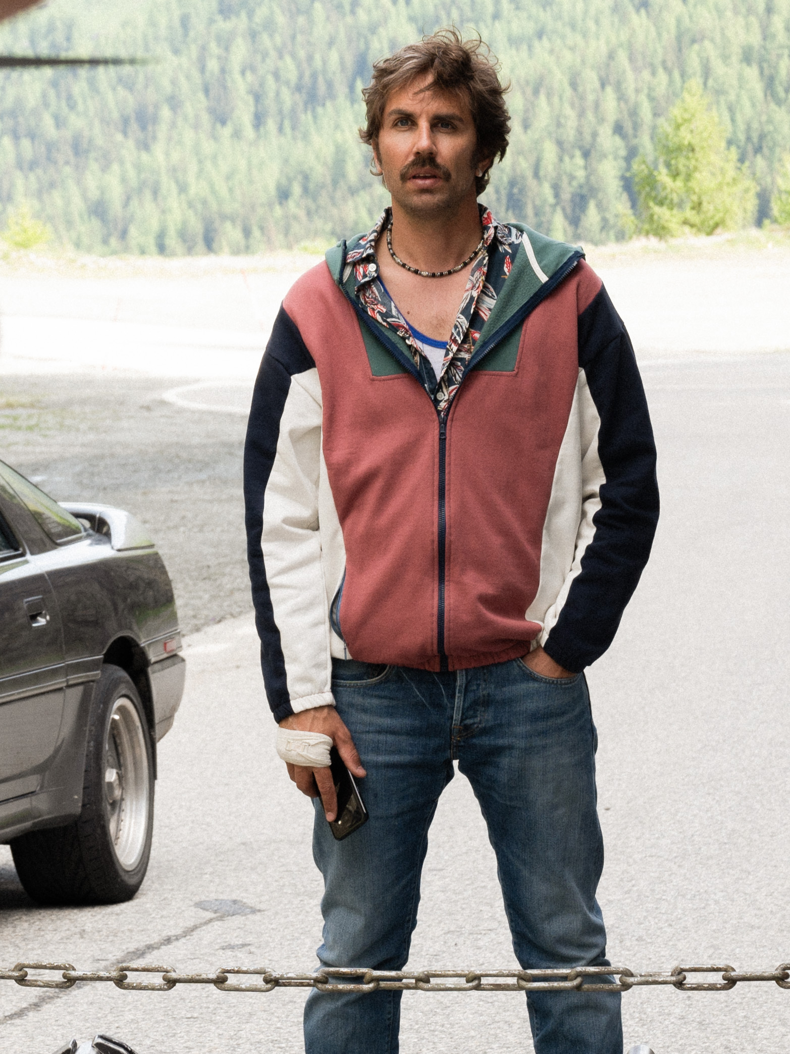 David Constantin as Bax stands in a red, white and green tracksuit beside a car on a mountain road in Tschugger.