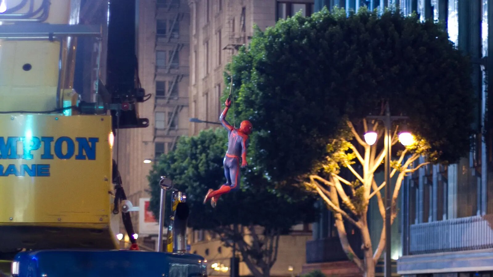 A Spider-Man stunt double swings on a wire above a night street while a camera crane and production vehicles frame the shot.