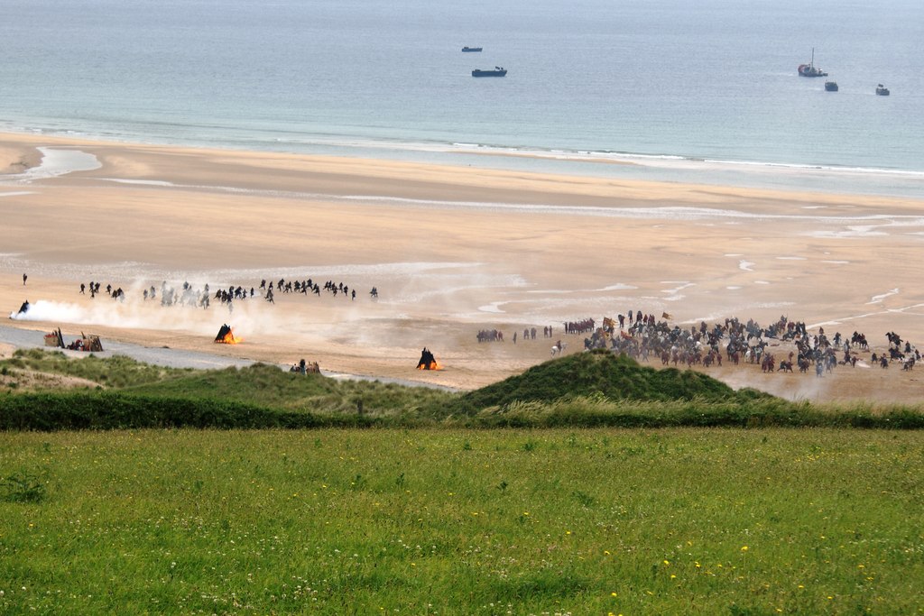 An elevated wide view of the Robin Hood battle shoot on Freshwater West beach shows large groups of performers, mounted riders, smoke, practical fires and boats offshore.