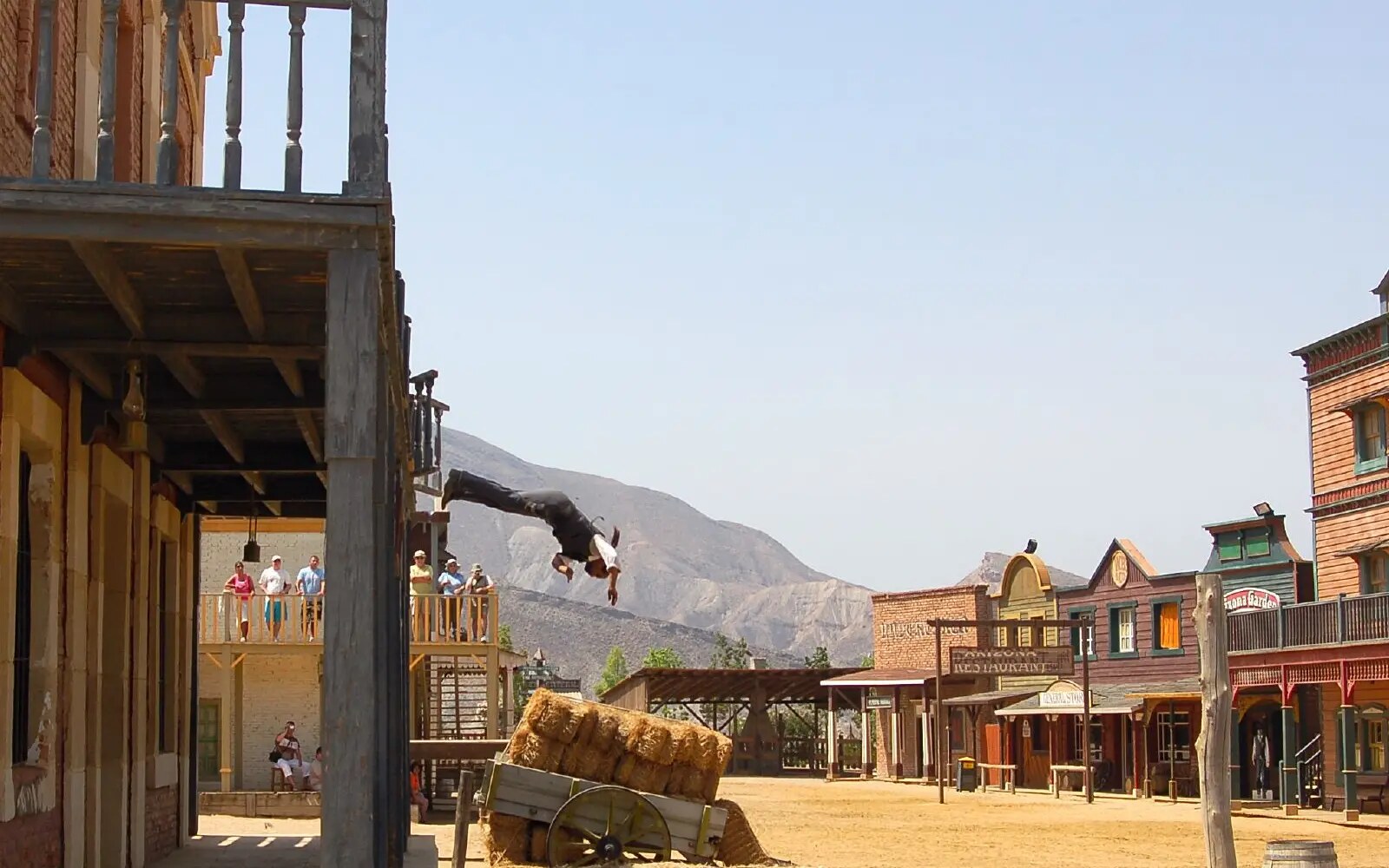 A stunt performer dives head-first from a Western-set balcony toward a hay-covered landing area.