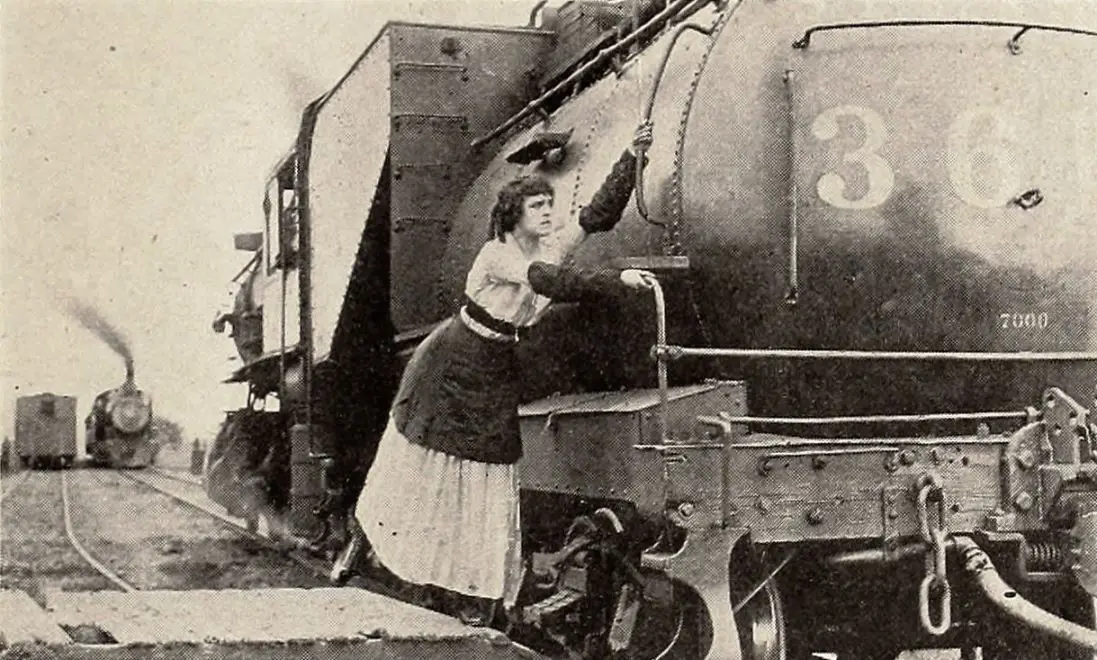 Helen Gibson stands on a locomotive's front side platform with one hand on a rail and the other on a rope in a June 1916 still from The Hazards of Helen.
