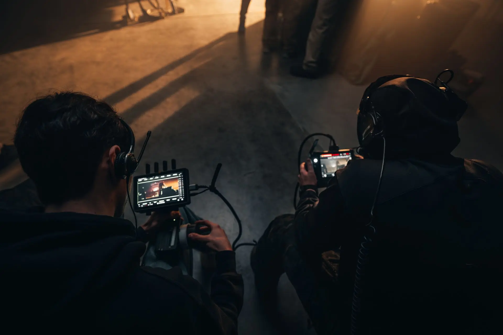 Two operators work side by side in a dark action set, one physically carrying a compact camera system and the other controlling the image from a separate monitor.