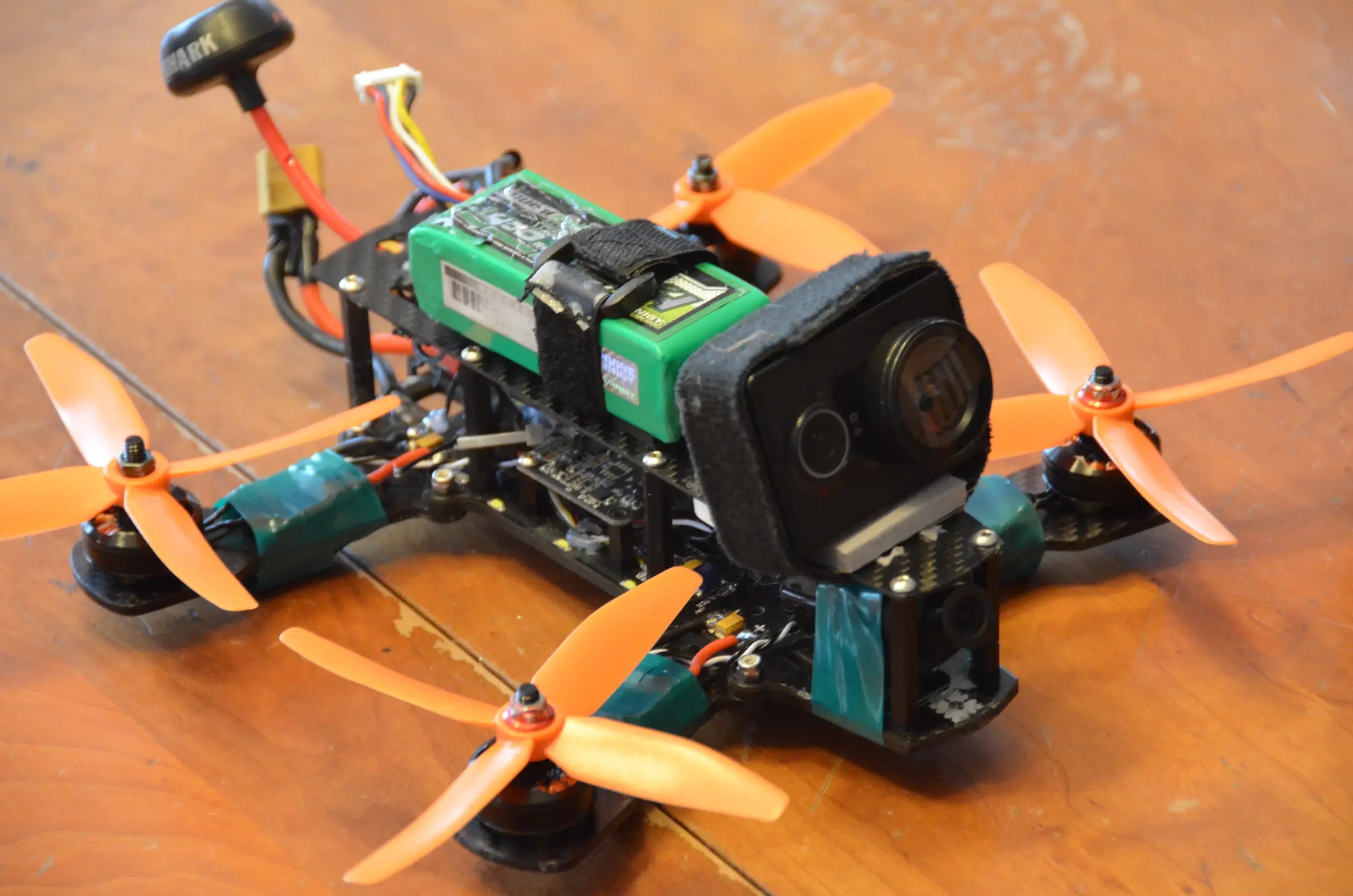 A compact racing FPV quadcopter with an onboard action camera, exposed electronics and four orange propellers.