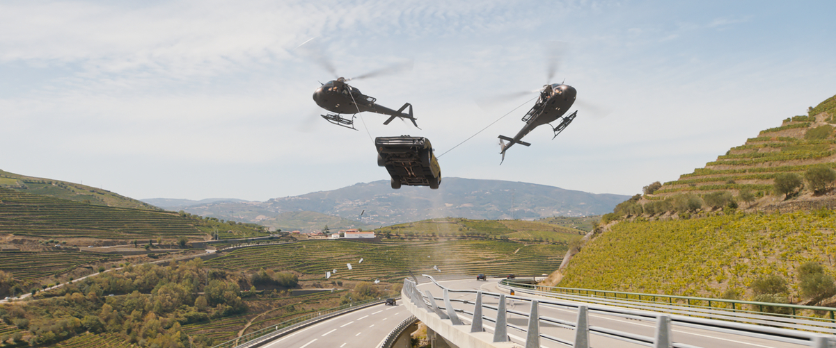 In a finished Fast X frame, two helicopters carry a black car above a curving mountain highway during the Portugal chase.
