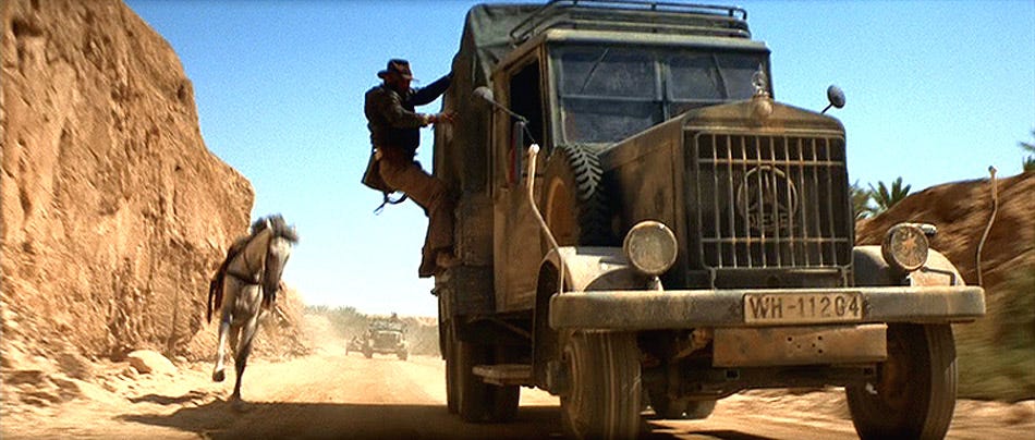 Indiana Jones clings to the side of a moving cargo truck while a white horse runs beside it during the desert chase in Raiders of the Lost Ark.