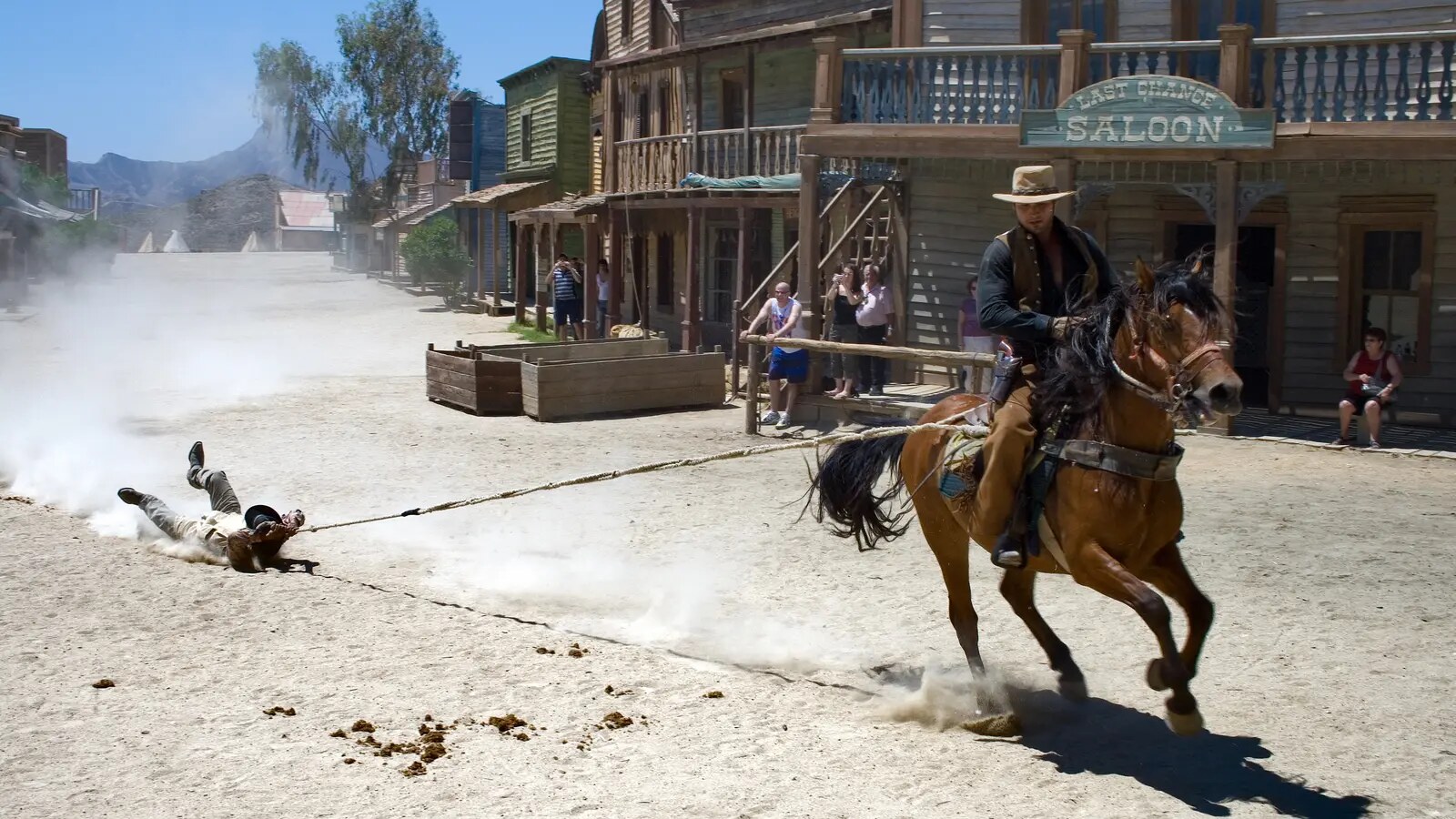 A stunt rider gallops a horse through a Western set while a second performer is dragged through dust on a controlled line behind.
