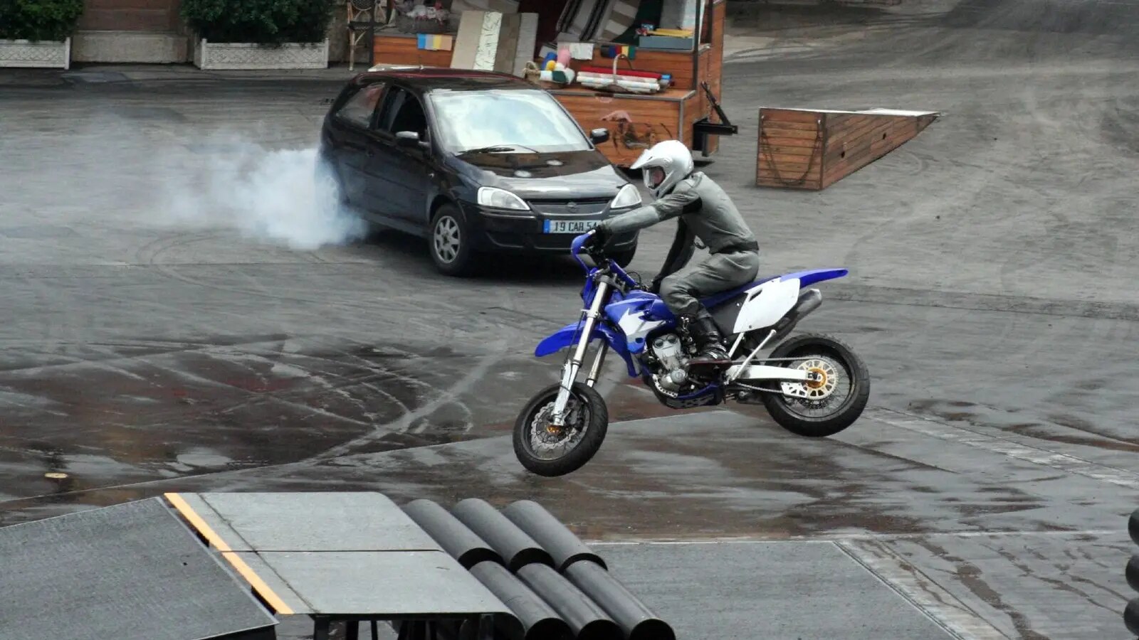 A stunt rider launches a motorcycle from a ramp as a car slides through tyre smoke behind the landing line.
