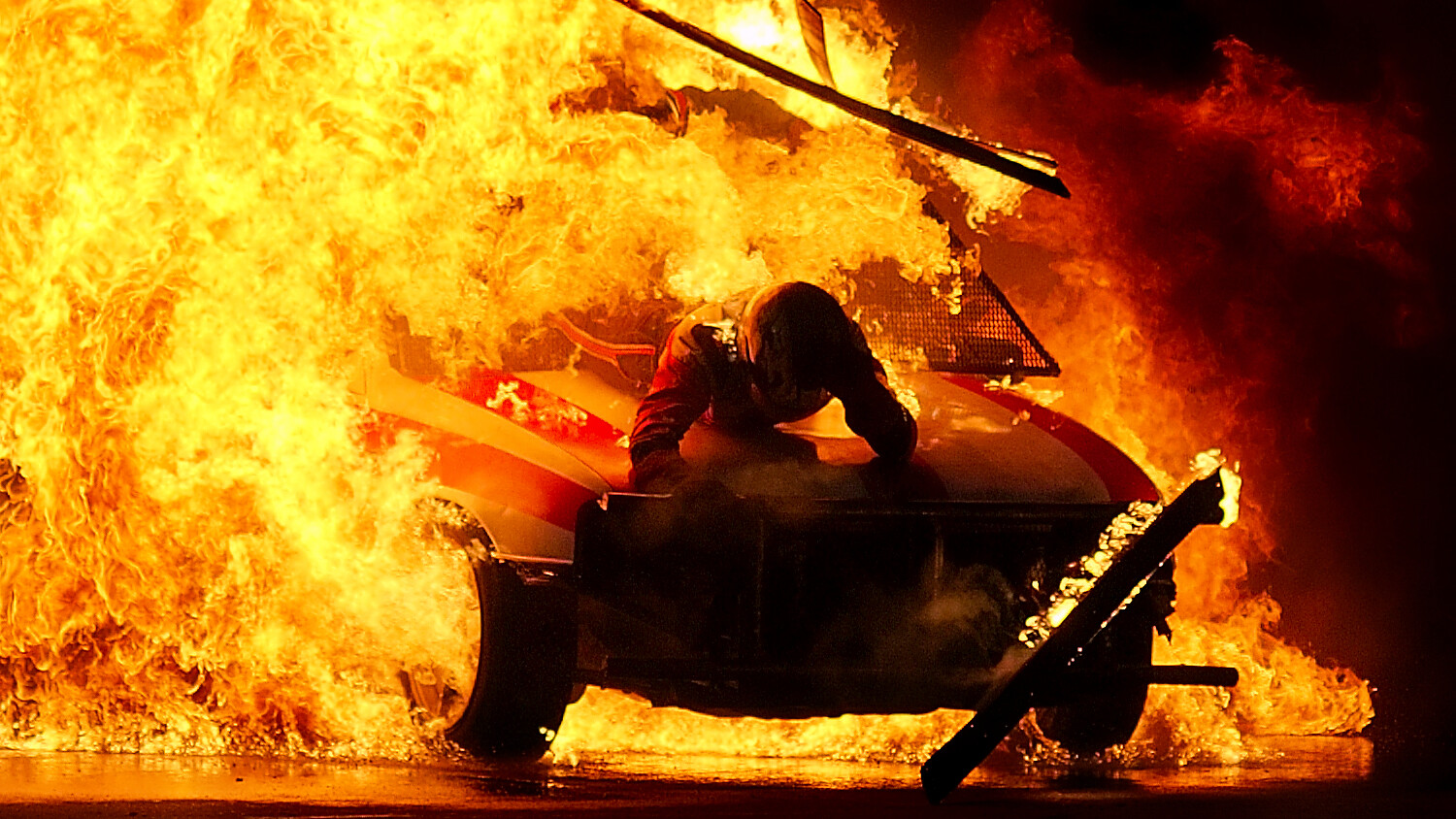 A stunt performer crouches on a car as a huge orange fireball fills the frame during a nighttime pyrotechnic exhibition.