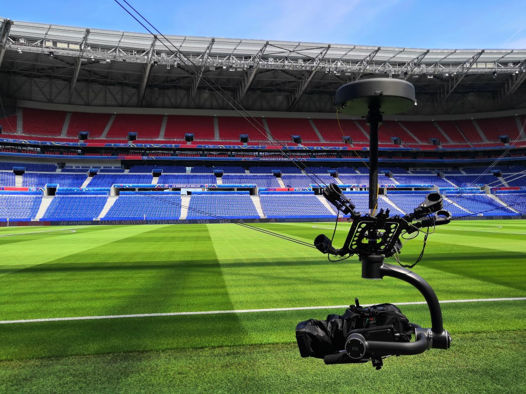 A cable-suspended Robycam camera head hangs above a football stadium in France, with multiple support lines visible.