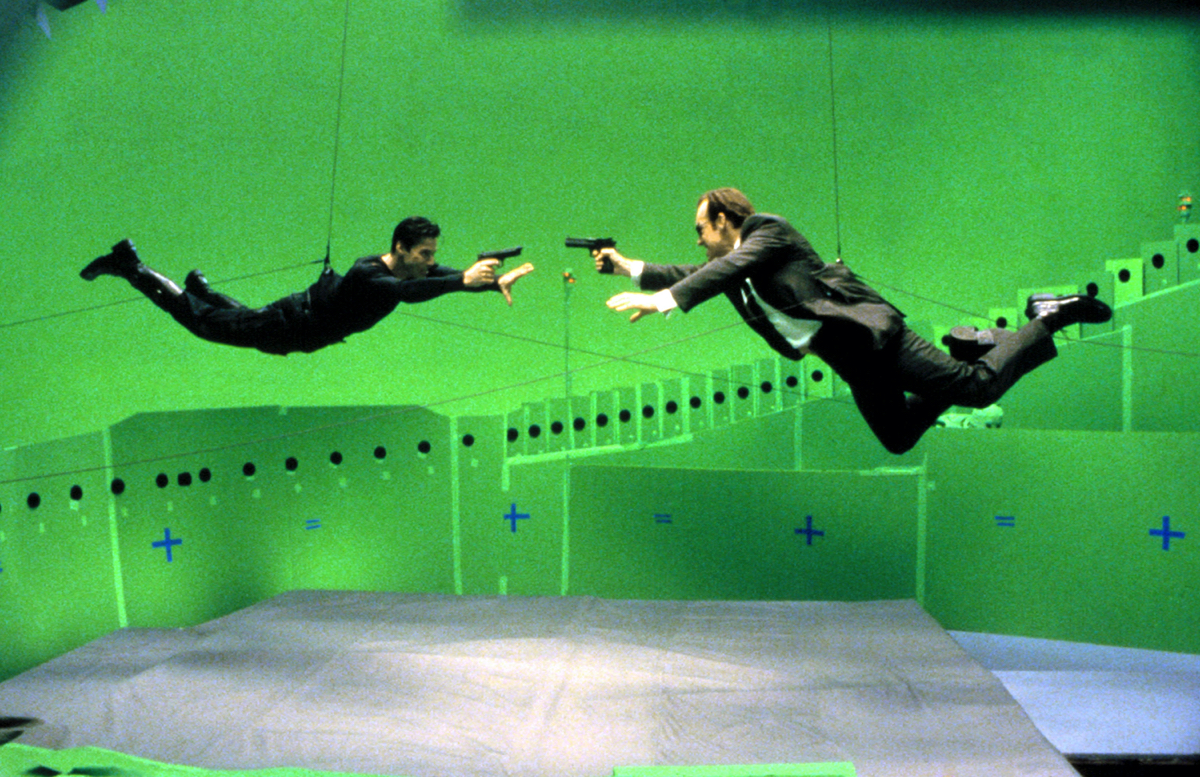 Two performers fly horizontally toward one another on visible wires above crash mats in a green-screen stage from The Matrix production.