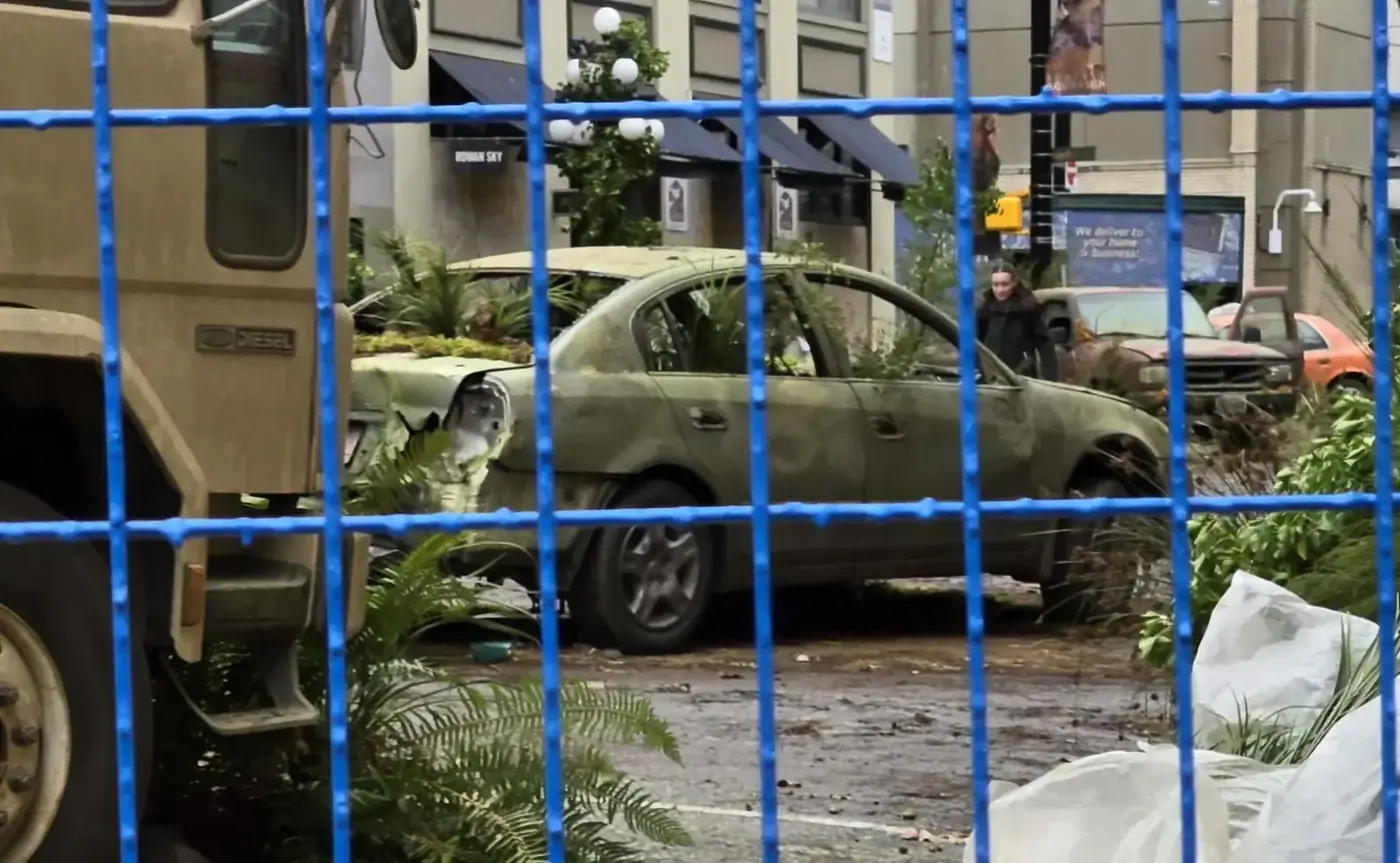 A blue perimeter fence separates the camera from a performer, weathered vehicles and overgrown street dressing on The Last of Us set in Vancouver.