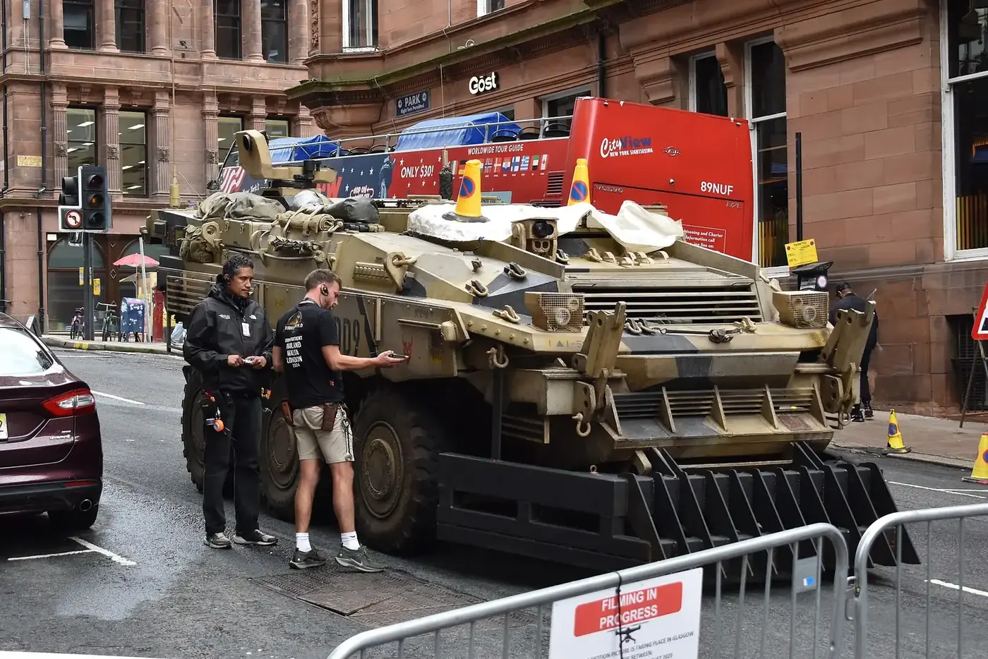 Two crew members stand beside a prop armoured vehicle behind filming barriers on a Glasgow street during production of Spider-Man: Brand New Day.
