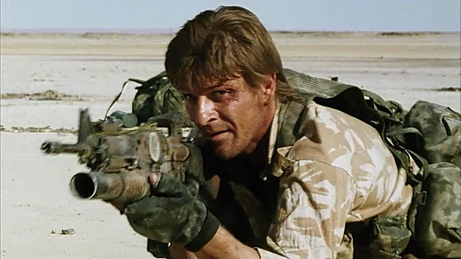 Sean Bean as Andy McNab crouches in an exposed pale desert with a rifle and heavy pack in Bravo Two Zero.