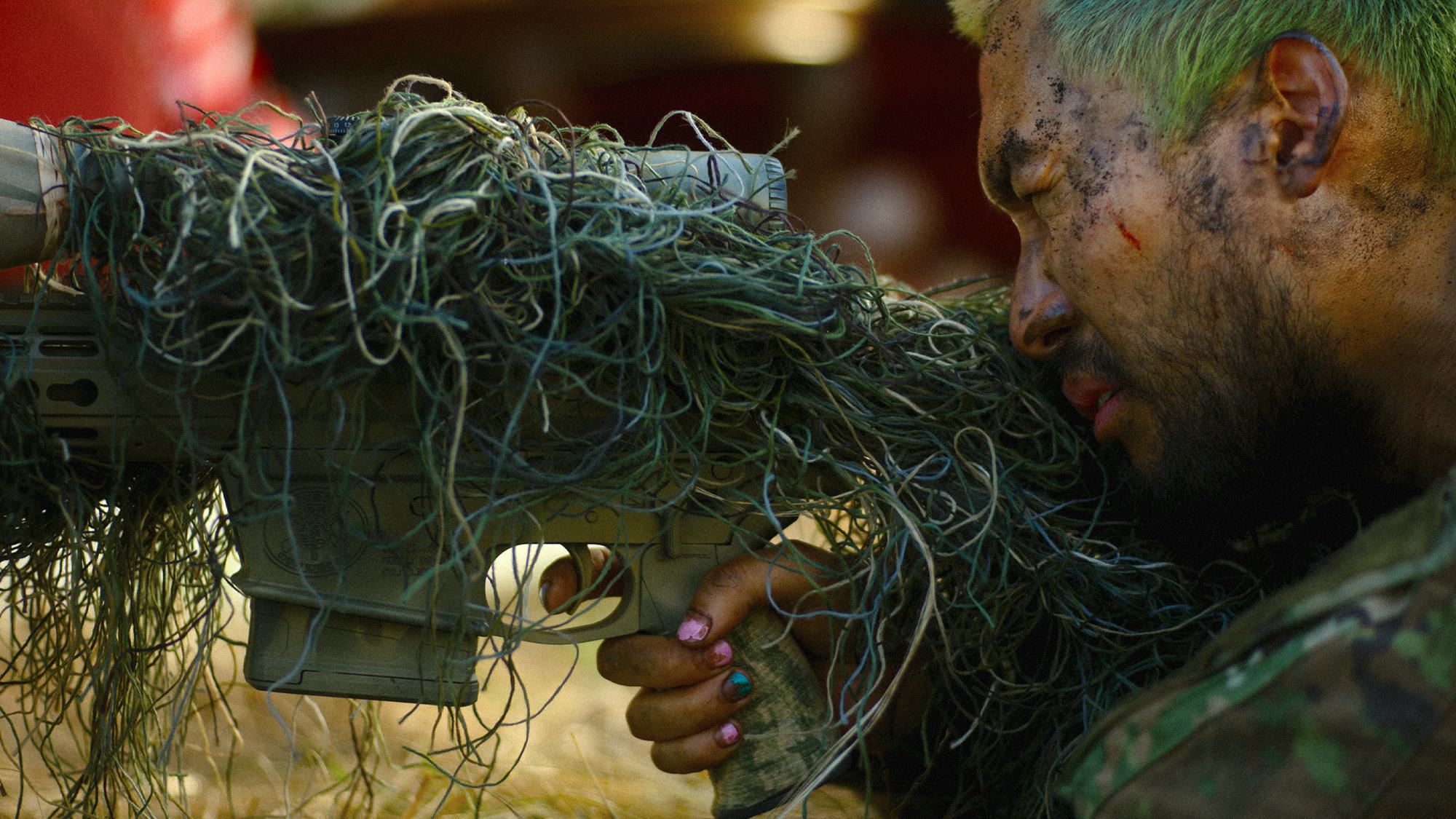 A mud-streaked combatant in Civil War leans against a rifle wrapped in camouflage fibres; brightly painted fingernails remain visible on the supporting hand.