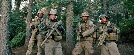Four members of the Lone Survivor cast stand close together with equipment and rifles among trees on uneven wooded ground.