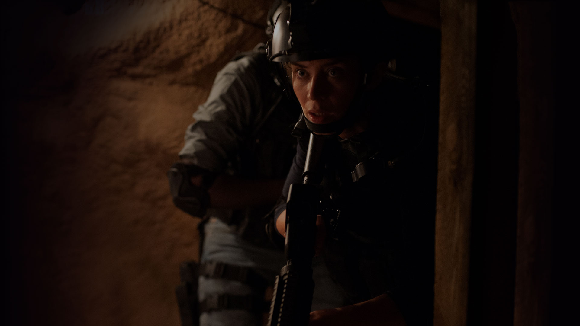 Emily Blunt as Kate Mercer holds position in a dark narrow passage in Sicario while a second figure remains partly obscured behind her.