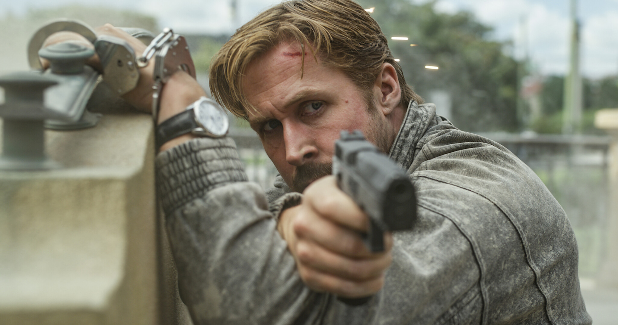 Ryan Gosling as Sierra Six, bloodied and handcuffed to a stone barrier, aims a pistol from cover during the Prague shootout in The Gray Man.