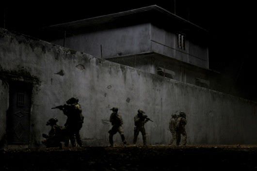 Five dark figures move along the exterior wall of the compound at night in Zero Dark Thirty, with only fragments of bodies and architecture visible.