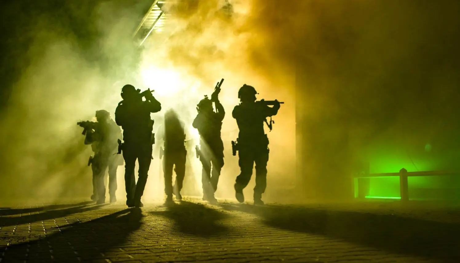 Four tactical performers move together out of foliage through green and yellow theatrical smoke during a staged outdoor production.
