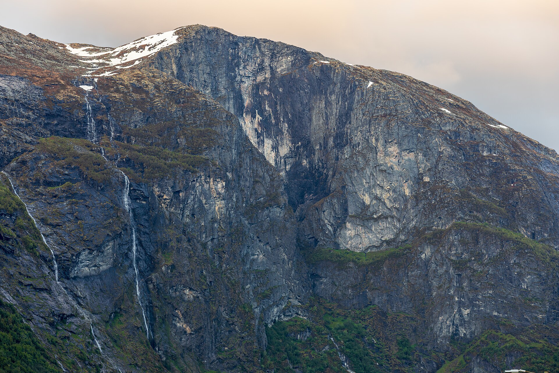 Die steile Felswand des Helsetkopen in Norwegen, mit Schneefeldern am Gipfel und schmalen Wasserfällen auf der linken Seite.